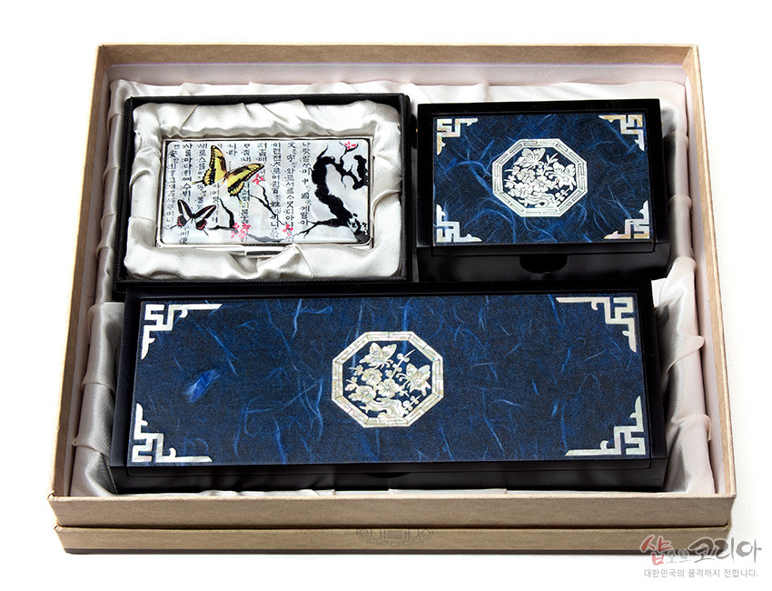 [텍스트: 샵오브코리아 대한민국의 품격까지 전달합니다.] 청색 한지 바탕 위 백색 자개 청매화와 나비 문양, 전통 기하학 테두리 장식이 조화를 이루는 필함·명함함·명함케이스 세트 이미지