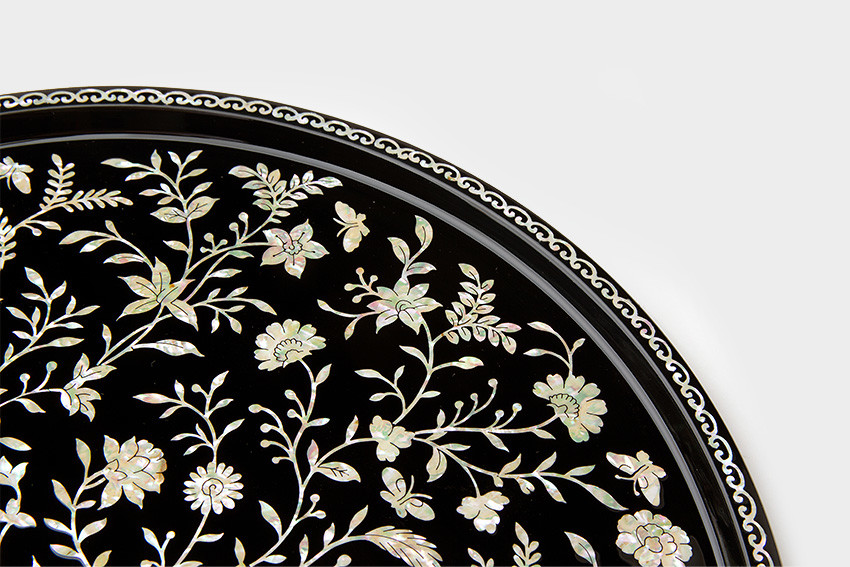 천년시 나전칠기 쟁반-나전넝쿨 검은 옻칠 바탕 위에 자개 상감으로 표현된 넝쿨과 꽃, 나비 문양이 섬세하게 펼쳐진 원형 나전칠기 쟁반 가장자리 파도무늬 장식 특징의 이미지 설명