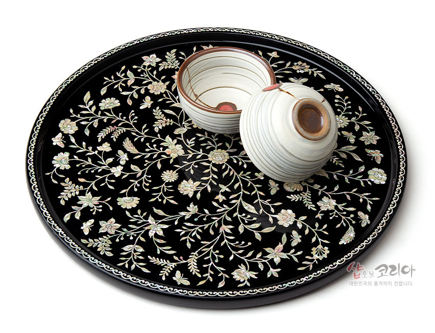 [텍스트: 샵오브코리아 대한민국의 품격까지 전합니다.] 짙은 검은 옻칠 바탕 위 자개 상감으로 넝쿨과 꽃문양을 촘촘히 배치한 원형 나전칠기 쟁반과 도자기 찻잔 배치 이미지 설명