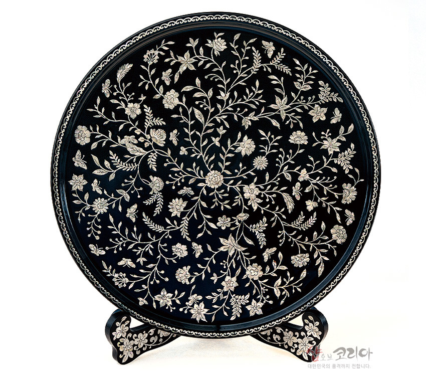 [텍스트: 샵오브코리아 대한민국의 품격까지 전합니다.] 짙은 흑색 옻칠 바탕 위 자개 상감으로 넝쿨과 꽃문양을 촘촘히 배치한 원형 나전칠기 쟁반과 곡선형 받침대 장식 소품