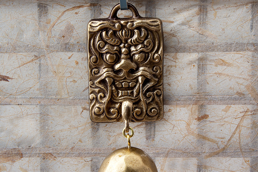 금속 재질의 직사각형 판에 입체적으로 새긴 도깨비 문양 장식, 하단에 매달린 황동빛 반구형 종이 어우러진 전통 풍경 디자인 이미지 설명