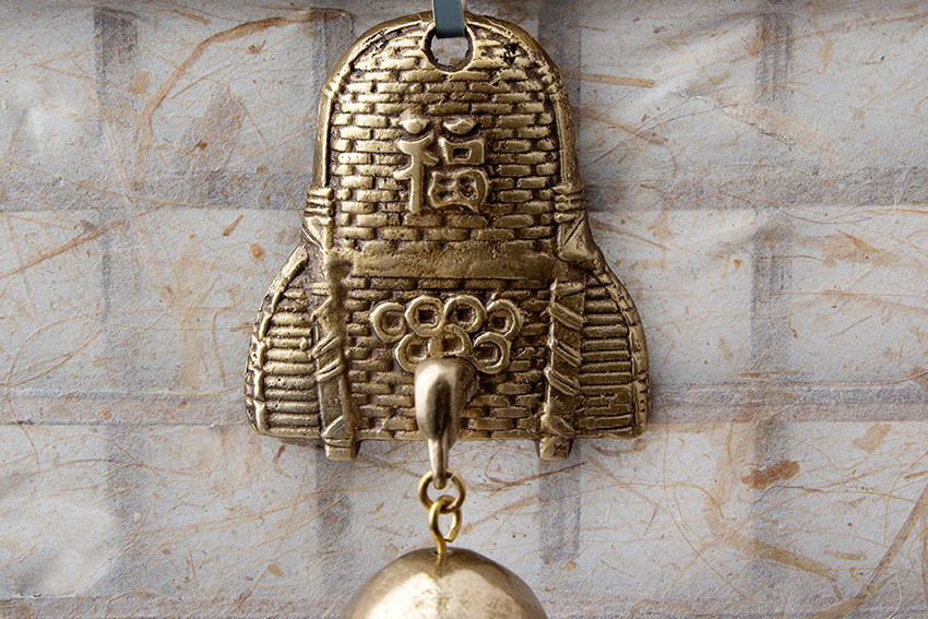 [텍스트: 福] 황동빛 금속 재질의 복주머니 형 문종 장식, 엿가락 매듭 문양과 양각 질감, 담백한 한지 느낌 배경 대비 구성