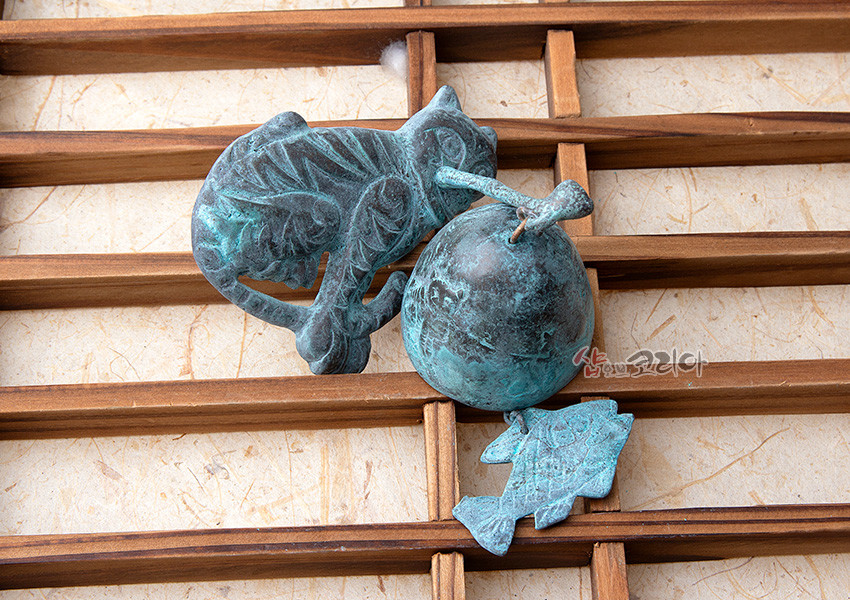 [텍스트: 샵오브코리아] 청동 재질에 청록색 산화 촉감을 살린 호랑이 형상 문종과 물고기 모양 추 장식, 나뭇결 나무 격자 위 배치된 이미지 묘사