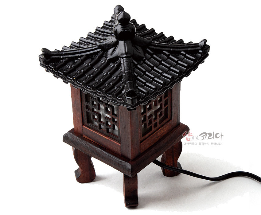 [텍스트: 샵오브코리아 대한민국의 품격까지 전합니다.] 짙은 갈색 목재와 흑색 기와 지붕으로 구성된 전통 정자형 미니 좌등, 완자문 창살과 반투명 창호지 느낌의 조명부가 있는 장식 조명소품 형상