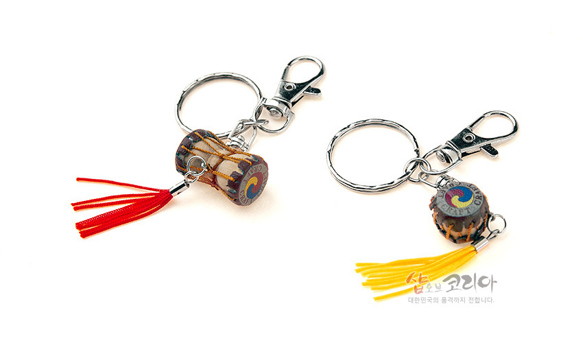 [텍스트: 샵오브코리아 대한민국의 품격까지 전합니다.] 금속 고리와 고리형 고정장식에 목재 질감 장구·북 미니어처, 태극 문양 포인트와 붉은색·노란색 술 장식 조합 이미지 설명