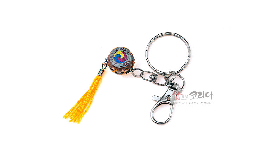 [텍스트: 샵오브코리아 대한민국의 품격까지 전합니다.] 금속 링과 고리, 노란 술 장식이 달린 목재 북 모양 장식, 상단에 삼색 태극 문양이 새겨진 열쇠고리 디자인