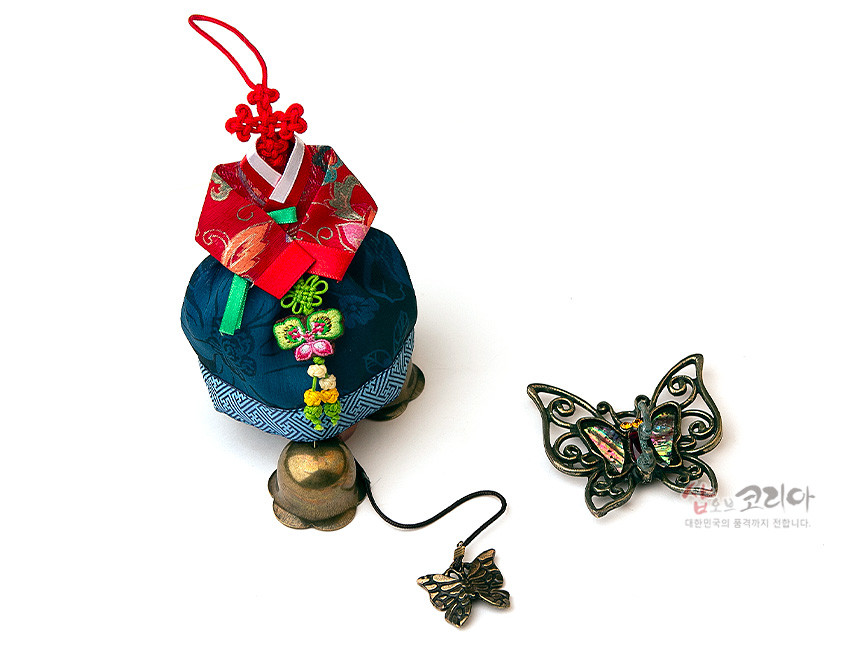 [텍스트: 샵오브코리아 대한민국의 품격까지 전합니다.] 적색 비단 저고리와 청색 비단 치마 한복 모양, 매듭 기법 장식과 수실 자수 문양, 청동색 금속 방울과 자개 장식 나비 형상 문종 구성