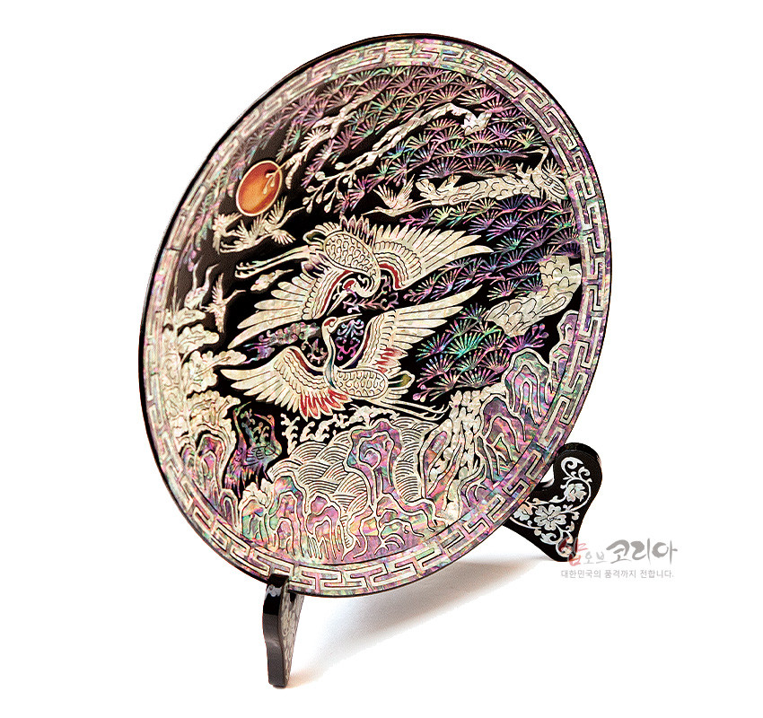 [텍스트: 샵오브코리아 대한민국의 품격까지 전합니다.] 흑색 옻칠 바탕에 자개 상감 기법으로 소나무와 쌍학, 일월·구름·산수 문양을 정교하게 표현한 원형 나전칠기 쟁반형 오브제