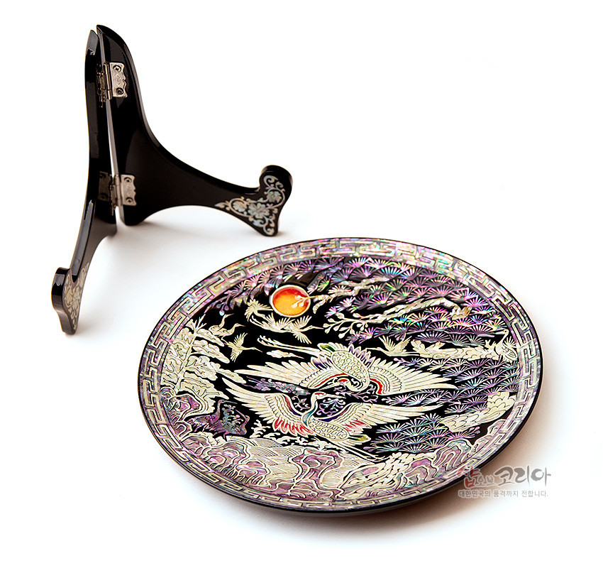 [텍스트: 샵오브코리아 대한민국의 품격까지 전합니다] 흑색 옻칠 바탕에 자개 상감으로 소나무와 해, 비상하는 쌍학을 표현한 원형 쟁반과 자개 장식 받침대 이미지 설명