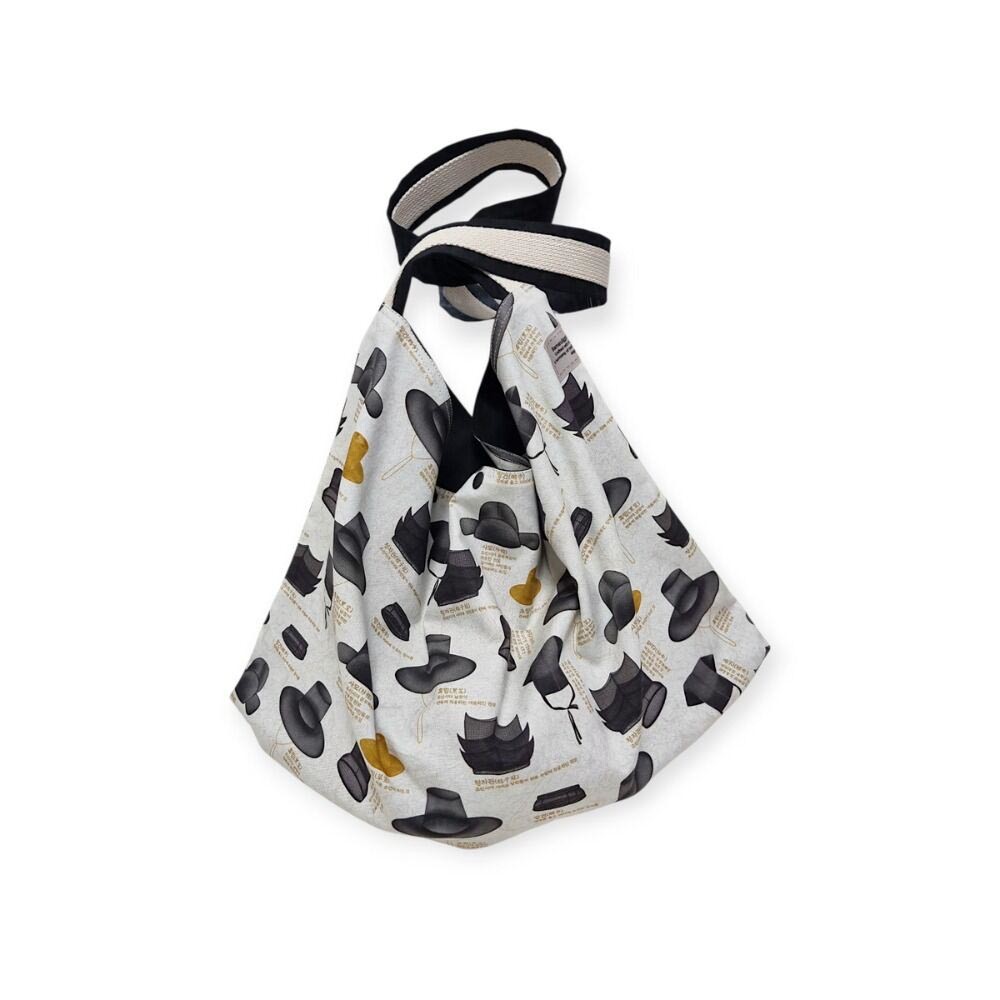 [텍스트: revele, 모자 관련 영문 문구] 전통 갓과 망건 일러스트가 반복된 패턴의 면 소재 에코백, 흑색·황토색 모티프와 아이보리 바탕의 대비가 특징인 디자인