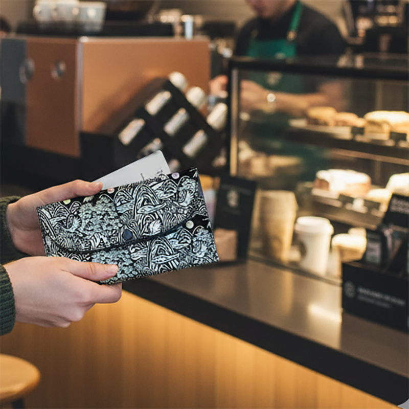 [텍스트: 없음] 카페 공간에서 사용되는 나전칠기 은빛 장생도 장지갑, 흑색 옻칠 위 자개 상감과 추상화된 장생도 문양의 대비가 돋보이는 디자인 연출