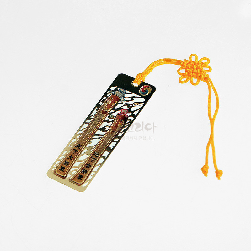 금속 재질에 금장 처리된 장승 문양과 구름 무늬, 노란 전통 매듭 술 장식이 있는 책갈피.