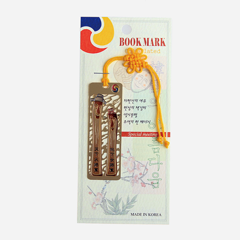 금속 재질에 장승 형상을 양각 처리하고 한글 문구를 새긴 책갈피와 노란 전통 매듭끈 장식.