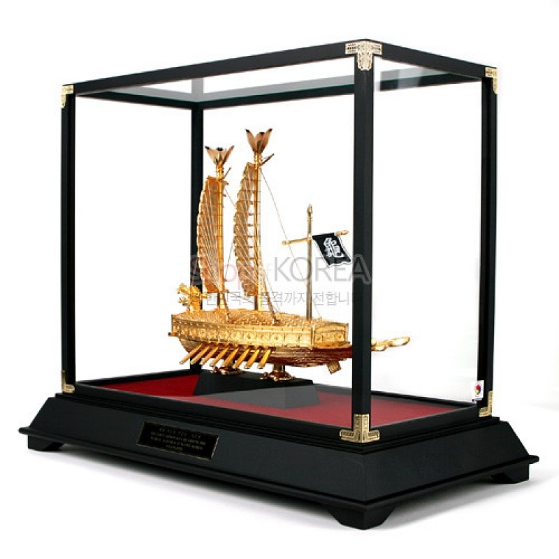 금빛 금속으로 세밀하게 재현된 거북선 모형과 유리 케이스, 검은 목재 받침이 조화된 장식품.