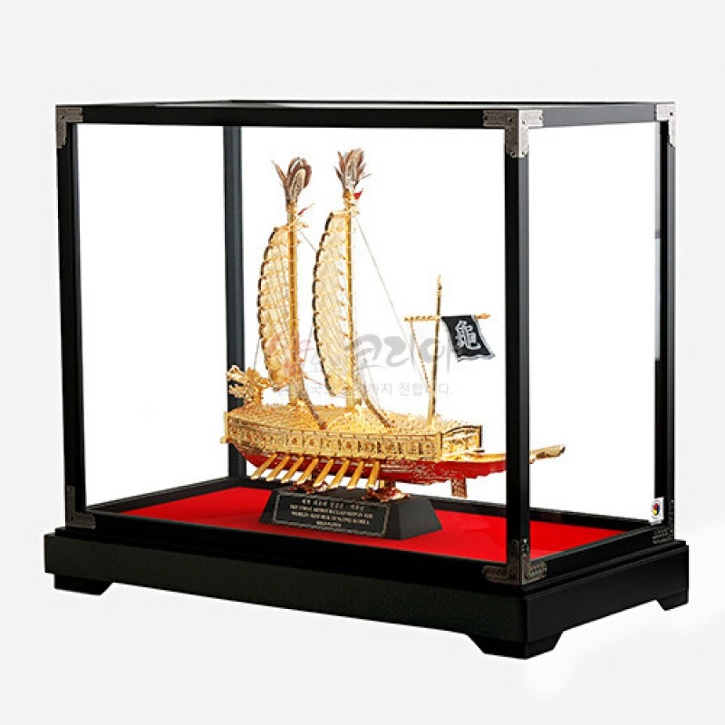 금속 재질의 금색 거북선 모형과 깃발 장식, 붉은 받침대와 유리 쇼케이스 구성이 특징인 장식품.