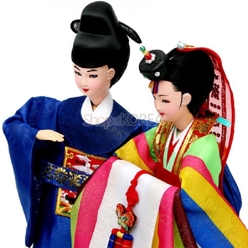 비단 재질의 전통 혼례 한복을 착용한 신랑신부 인형, 금박 장식과 오방색 색동, 매듭 노리개 장식 특징.
