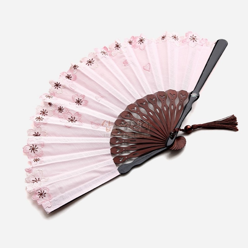 연분홍색 천에 매화 자수 문양이 수놓인 비단 접이부채, 나뭇결 살대와 매듭 장식 술이 어우러진 디자인.