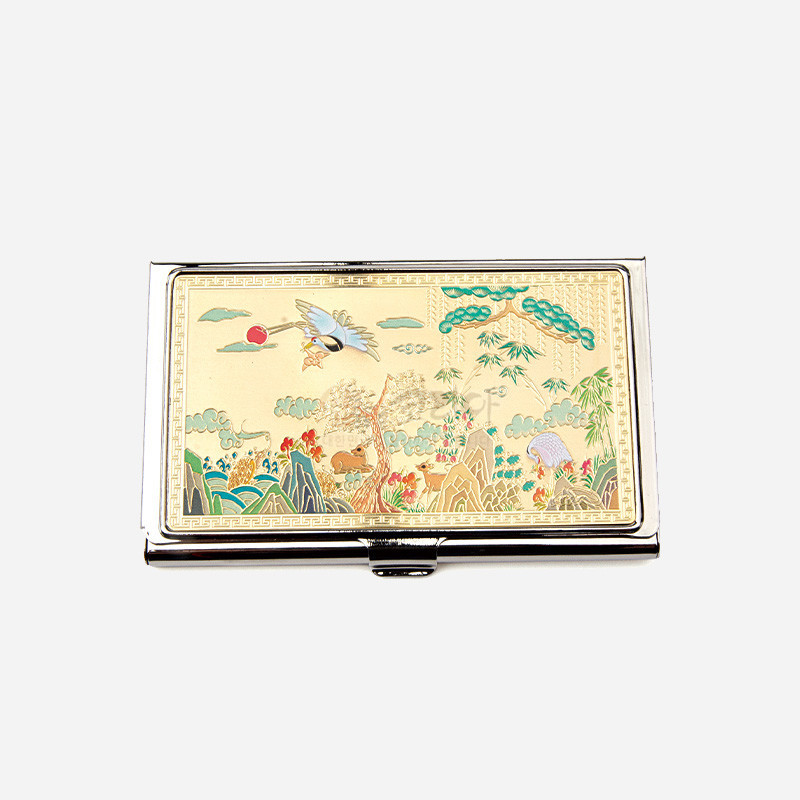 금속 소재 위에 금장 처리와 채색 기법으로 장생도 산수와 학, 구름, 대나무 문양을 섬세하게 표현한 명함케이스.
