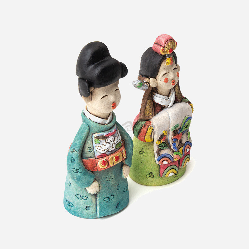 채색 도자 재질로 제작된 한복 차림 신랑신부 인형, 청록·연두색과 다채로운 자수 문양이 특징인 장식품.