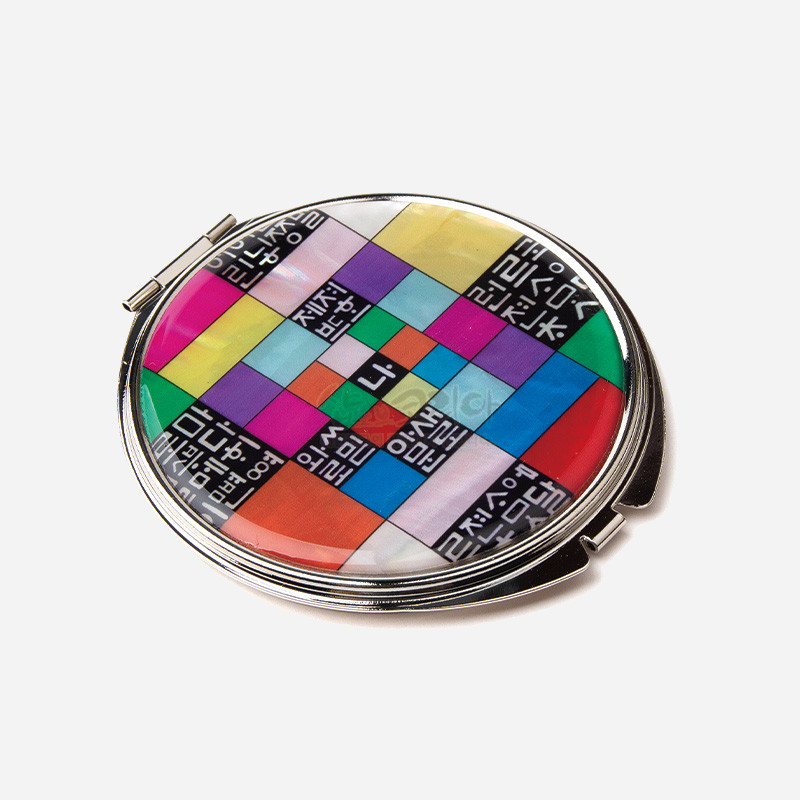 금속 테두리에 자개 느낌의 광택을 입히고, 오방색 조각보와 흑백 훈민정음 글자 문양이 어우러진 원형 손거울.