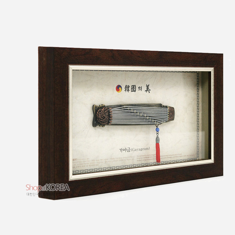 나무 프레임 속 금속 가야금 모형과 붉은 술 장식, 담백한 한지 배경과 전통 문양 테두리가 조화를 이룬 액자형 장식.