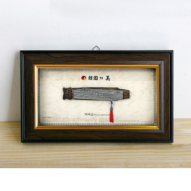 나무 느낌의 액자 속에 금속 재질 가야금 모형과 전통 무늬 매듭, 붉은 술 장식이 어우러진 장식 액자.