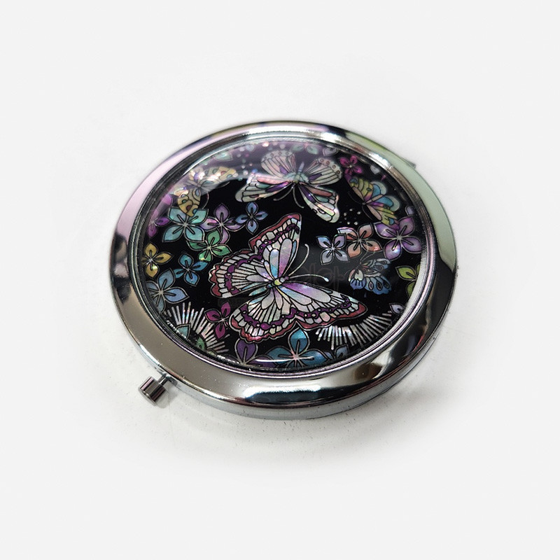 검은 바탕 위에 자개 상감 기법으로 표현된 꽃과 나비 문양이 돋보이는 금속 테두리 손거울.