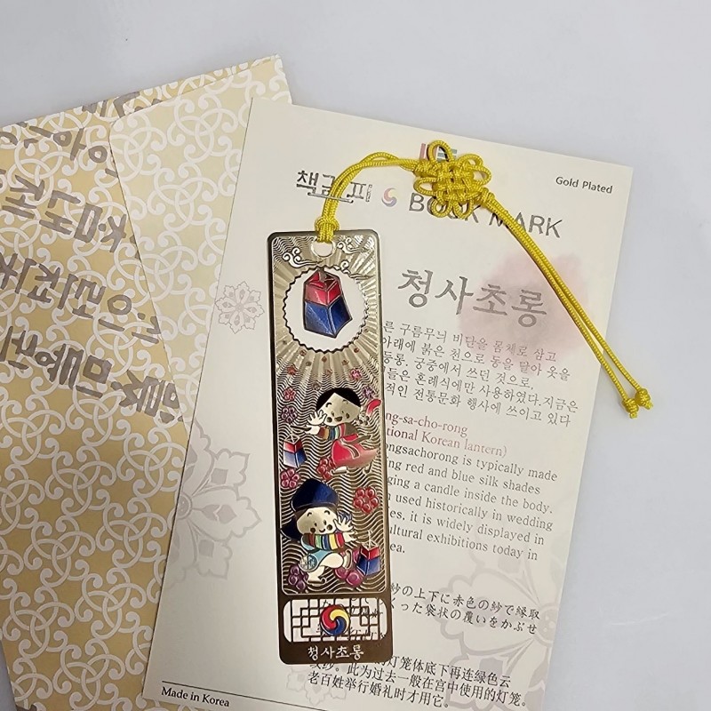 금장 금속 소재에 청사초롱과 한복 입은 아이들 문양, 다채로운 채색과 노란 매듭 끈 장식이 특징인 책갈피.