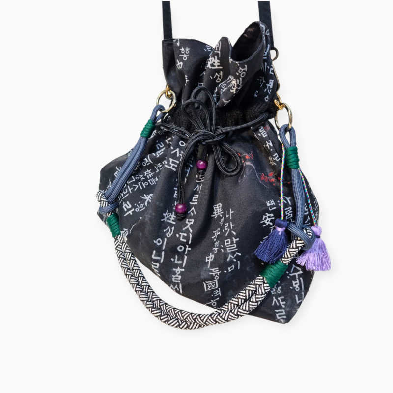 검은 비단 소재에 훈민정음 서체 문양이 인쇄된 버킷백, 매듭 장식과 다색 술, 전통 패턴 끈이 어우러진 디자인.