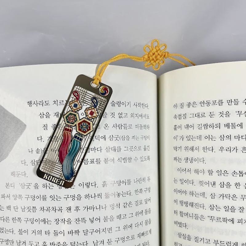 금속 재질 위에 칠보 기법으로 표현된 오방색 노리개 문양과 황색 끈 매듭 장식이 조화를 이룬 책갈피.