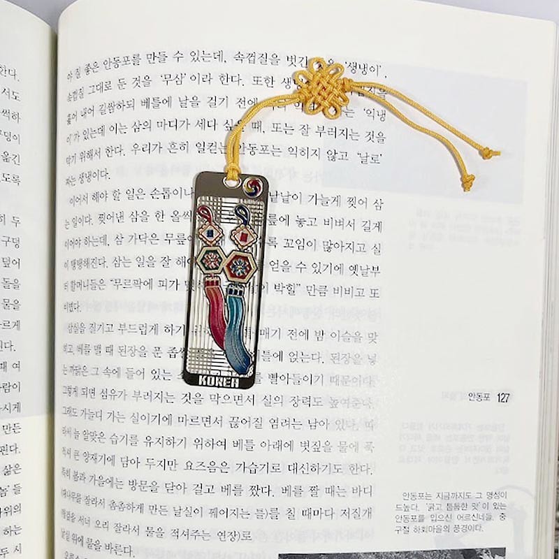금속 소재에 노리개와 태슬 문양을 레이저 절삭으로 표현하고 노란색 전통 매듭 끈을 더한 직사각형 책갈피.
