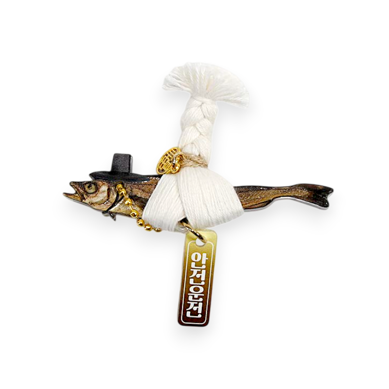 금속 재질의 명태 형상 위에 흰색 끈 매듭과 술 장식을 올리고, 금색 금속 장식과 한글 각인 패턴이 더해진 차량 송풍구 액세서리.