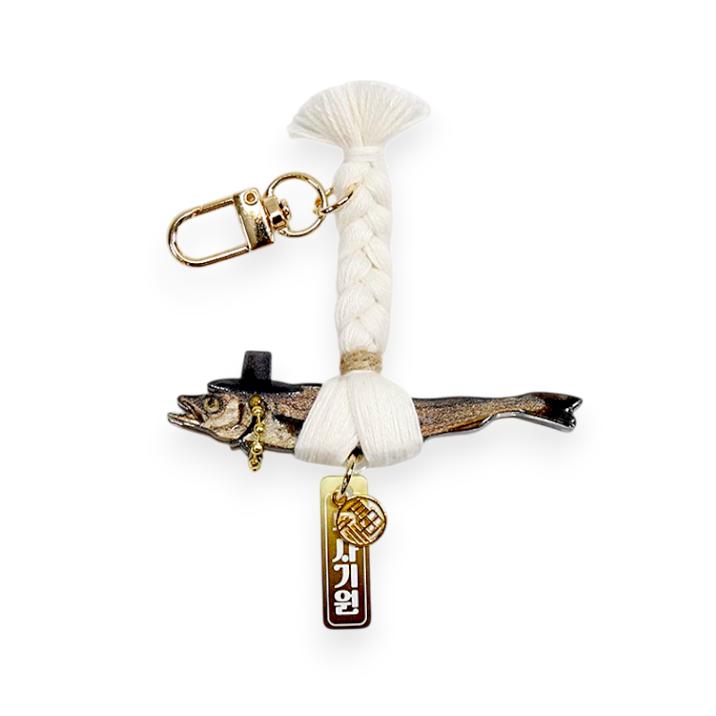 금속 소재 명태 모양 장식에 흰색 실 땋기 매듭과 금색 금속 고리, 문자 음각 펜던트가 어우러진 열쇠고리형 장식품.