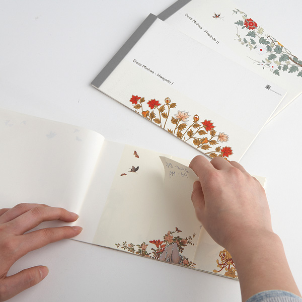 [텍스트: Damt hithra - Hwajodo 1] 흰색 종이 재질에 회색 제본 처리, 하단에 붉은 꽃과 연한 갈색 잎사귀가 조화된 화조도 민화 문양, 펼침면 하단에 꽃과 새, 나비가 섬세하게 인쇄된 메모지 디자인