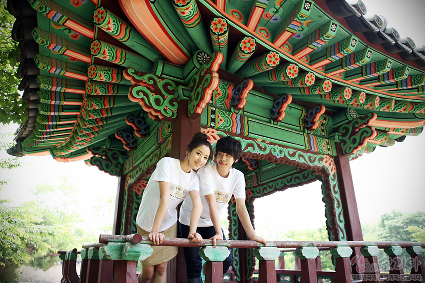 [텍스트: SHOP OF KOREA] 단청 문양으로 장식된 전통 정자와 흰색 면 소재 3D 입체 발포 도깨비 티셔츠 착용 남녀 모델 연출 이미지