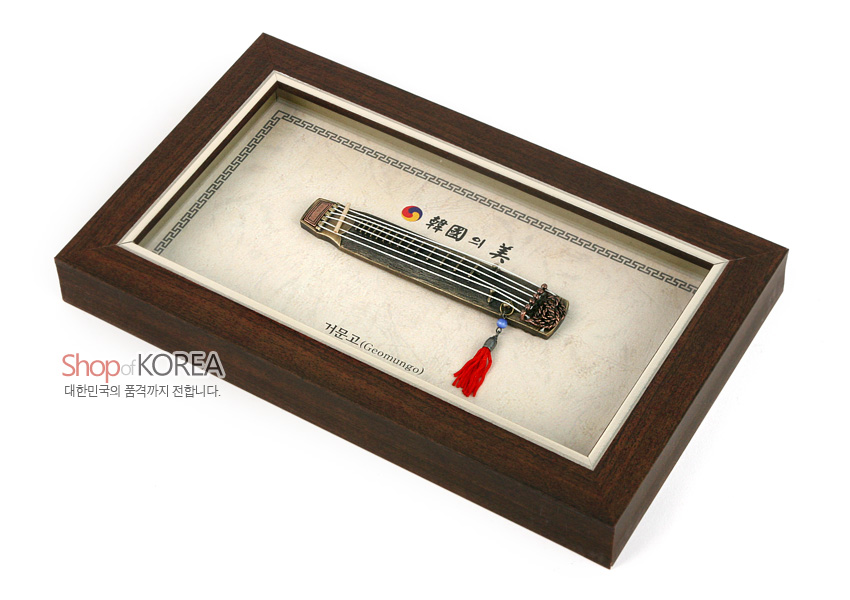[텍스트: Shop of KOREA / 대한민국의 품격까지 전합니다. / 韓國의 美 / 가야고 (Geomungo)] 짙은 갈색 나무 질감 액자 속 금속 재질 거문고 모형과 태극 문양, 붉은 술 장식이 조화를 이룬 전통 악기 장식 액자