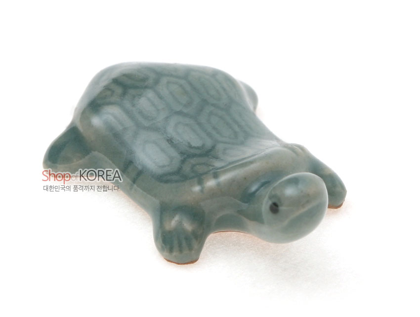 [텍스트: Shop of KOREA 대한민국의 품격까지 전달합니다.] 옅은 비취색 청자 재질의 거북이 형 수저받침, 육각형 등껍질 문양과 매끈한 유약 광택이 돋보이는 도자 공예품 형상