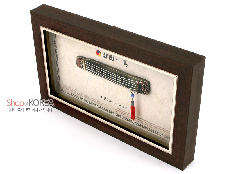 [텍스트: Shop of KOREA, 대한민국의 품격까지 전합니다. 韓國의 美, 거문고(Geomungo)] 짙은 갈색 원목 느낌 액자 속 금속 재질 미니어처 거문고와 태극 문양, 청색·적색 술 장식, 전통 문양 테두리 디자인 이미지 설명