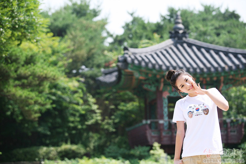 [텍스트: KOREA] 흰색 면 티셔츠 위 태극 문양과 전통 탈 그래픽, 한글 서체가 조화된 3D 발포 프린트 장식, 배경 전통 누각과 녹음 요소 이미지