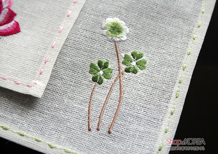 [텍스트: ShopofKOREA 대한민국의 품격까지 전합니다] 모시 천 위에 흰꽃과 초록 잎 자수, 가장자리에 분홍·연두 스티치로 장식한 전통 식탁보 디테일 표현