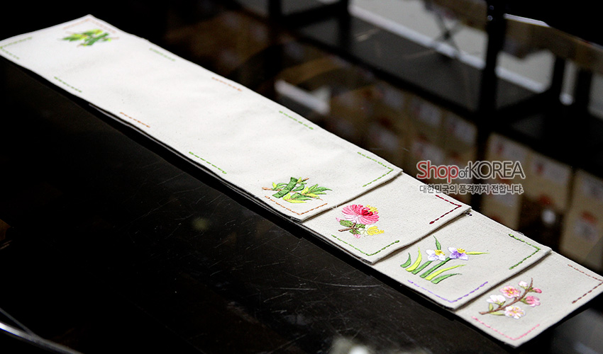 [텍스트: Shop of KOREA 대한민국의 품격까지 전합니다] 광목 재질 긴 직사각형 식탁보, 가장자리 컬러 스티치와 끝부분 대나무·국화·난초·매화 자수 문양 장식