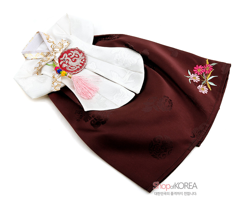 [텍스트: Shop of KOREA / 대한민국의 품격까지 전합니다.] 갈색 비단 치마와 은은한 무늬의 흰색 저고리 구성, 꽃문양 자수와 분홍 술 노리개 매듭 장식의 당의 형식 와인 커버 이미지 설명