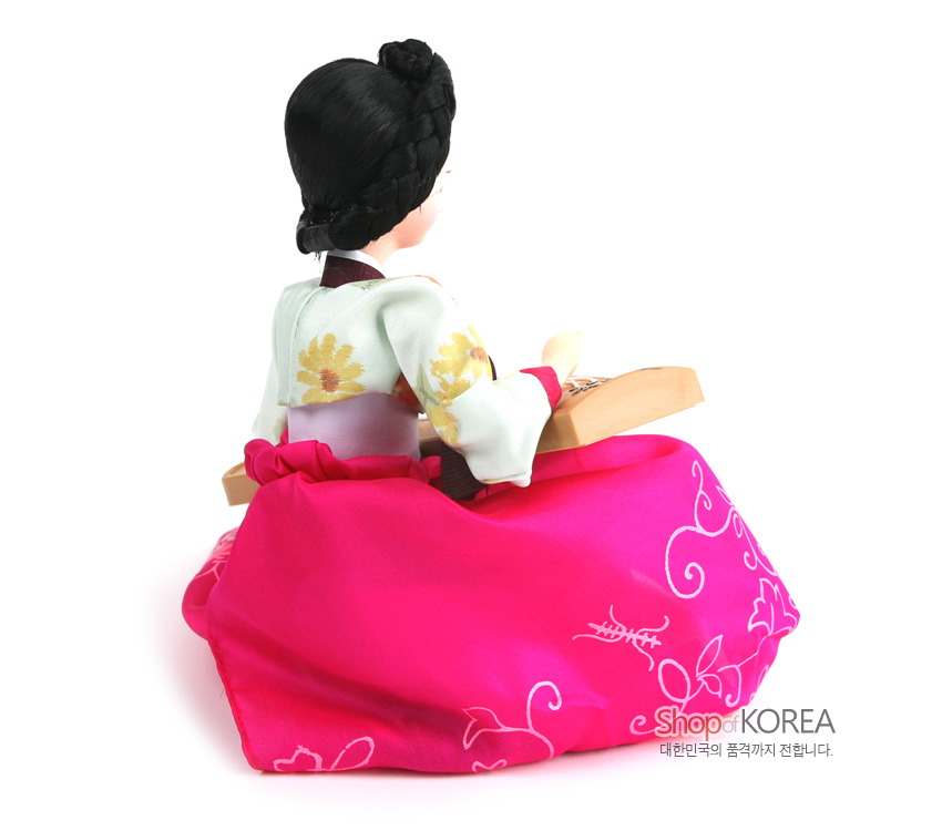 [텍스트: Shop of KOREA 대한민국의 품격까지 전합니다.] 분홍 비단 치마에 흰 나염 꽃무늬 장식, 연두 저고리에 노란 꽃문양 직조, 목재 가야금을 안고 앉은 전통 한복 인형 뒷모습 묘사