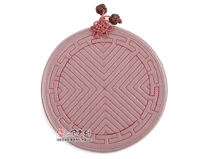 [텍스트: 샵오브코리아 대한민국의 품격까지 전합니다.] 파스텔 핑크 톤 천 소재에 전통 누비 기법으로 기하학 문양을 넣고 상단에 장식 매듭을 더한 원형 컵받침 이미지 묘사