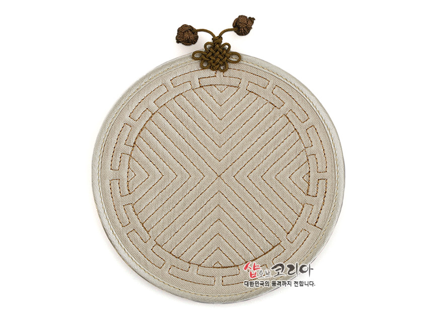 [텍스트: 샵오브코리아 대한민국의 품격까지 전합니다.] 아이보리 톤 천 위 전통 누비 기법으로 기하학 문양을 수놓고 상단에 갈색 배씨머리 매듭 장식을 더한 원형 컵받침 디자인