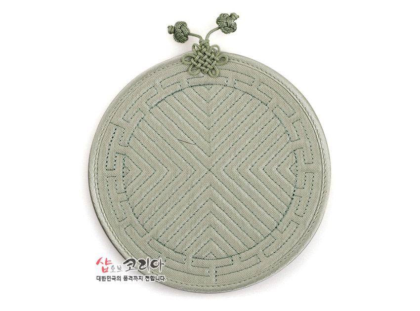 [텍스트: 샵오브코리아 대한민국의 품격까지 전합니다.] 연녹색 원형 천 소재에 전통 누비 기법으로 사선 기하학 문양을 넣고 상단에 매듭 장식을 더한 컵받침 이미지 설명