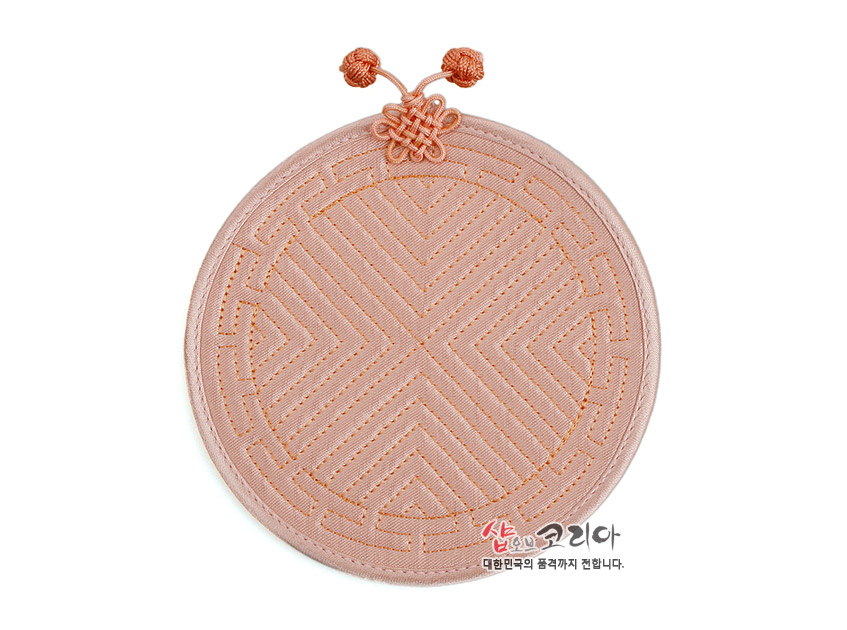 [텍스트: 샵오브코리아 대한민국의 품격까지 전합니다.] 파스텔 살구색 원형 천 소재에 전통 누비 기법으로 기하학 문양을 넣고 상단 배씨머리 매듭 장식을 더한 컵받침 모습