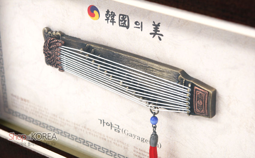 [텍스트: 韓國의 美, 가야금(Gayageum), ShopofKOREA] 금속 재질 가야금 모형과 흰색 줄 장식, 청색 구슬과 붉은 술이 어우러진 전통 문양 액자 구성
