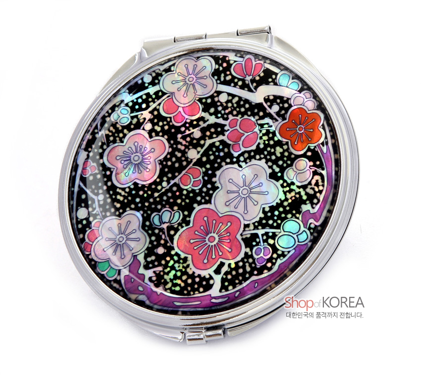 [텍스트: Shop of KOREA 대한민국의 품격까지 전합니다.] 금속 테두리 안 흑색 바탕에 자개 상감 기법으로 표현된 다채로운 색매화와 가지 문양이 화려하게 펼쳐진 원형 손거울 디자인