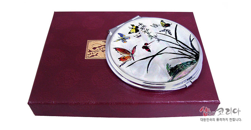 [텍스트: 샵오브코리아 대한민국의 품격까지 전합니다.] 자개 바탕 위 옻칠 기법으로 표현된 난초와 다색 나비 문양의 원형 금속 손거울과 붉은 비단 문양 선물 상자 조합 이미지